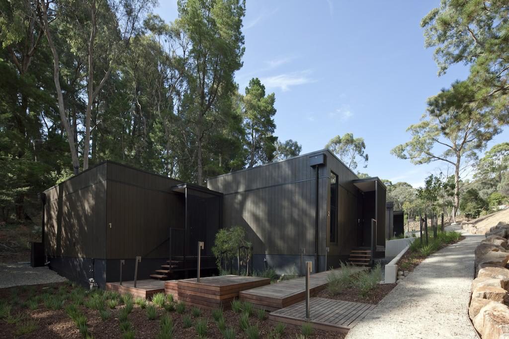 Outdoor view of Villas 1 and 2 framed by the native Australian flora and lush Hepburn gardens to the rear of the villas, Geometric wooden paths lead to private Villa entrances and a path leading to guest parking is lined with neutral local standstone boulders. View from outside reception building facing easterly mid morning.